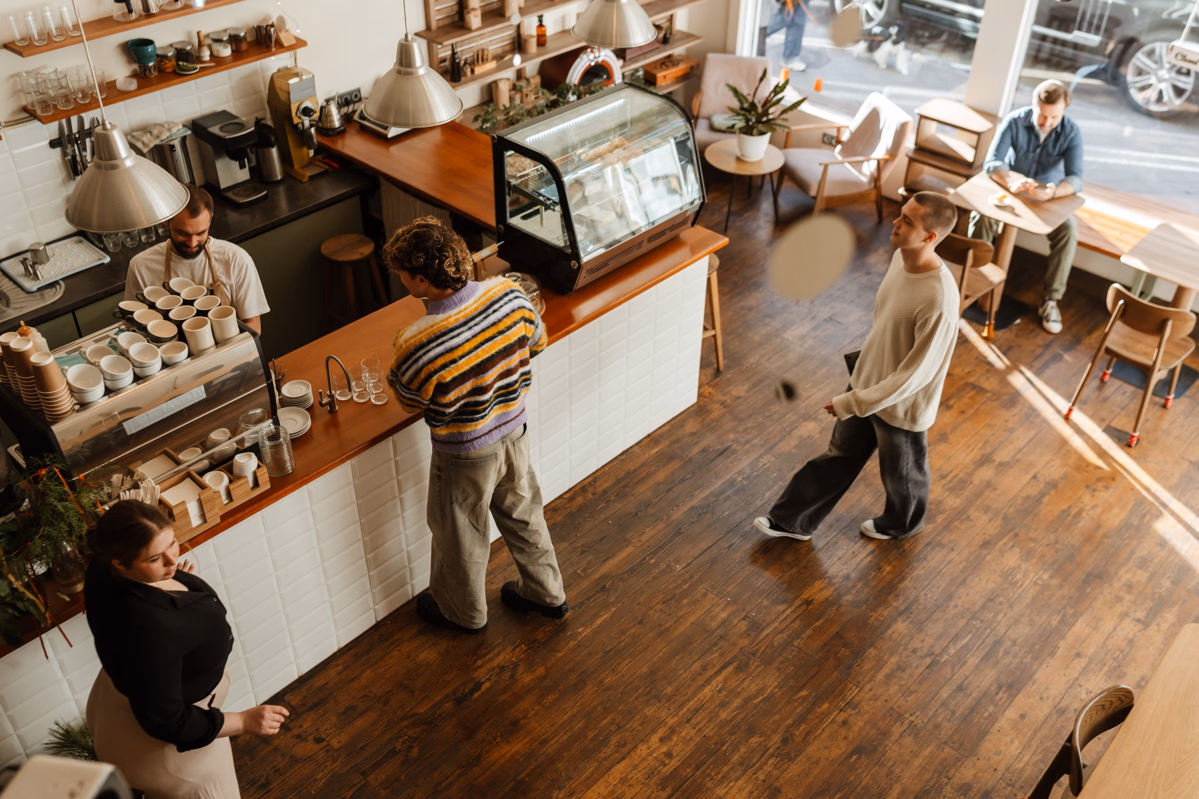Blick von oben in ein Café: Ein Gast wartet an der Theke, der Barista stellt Tassen bereit, ein weiterer Gast kommt herein, am Fenster sitzt ein Mann mit Smartphone
