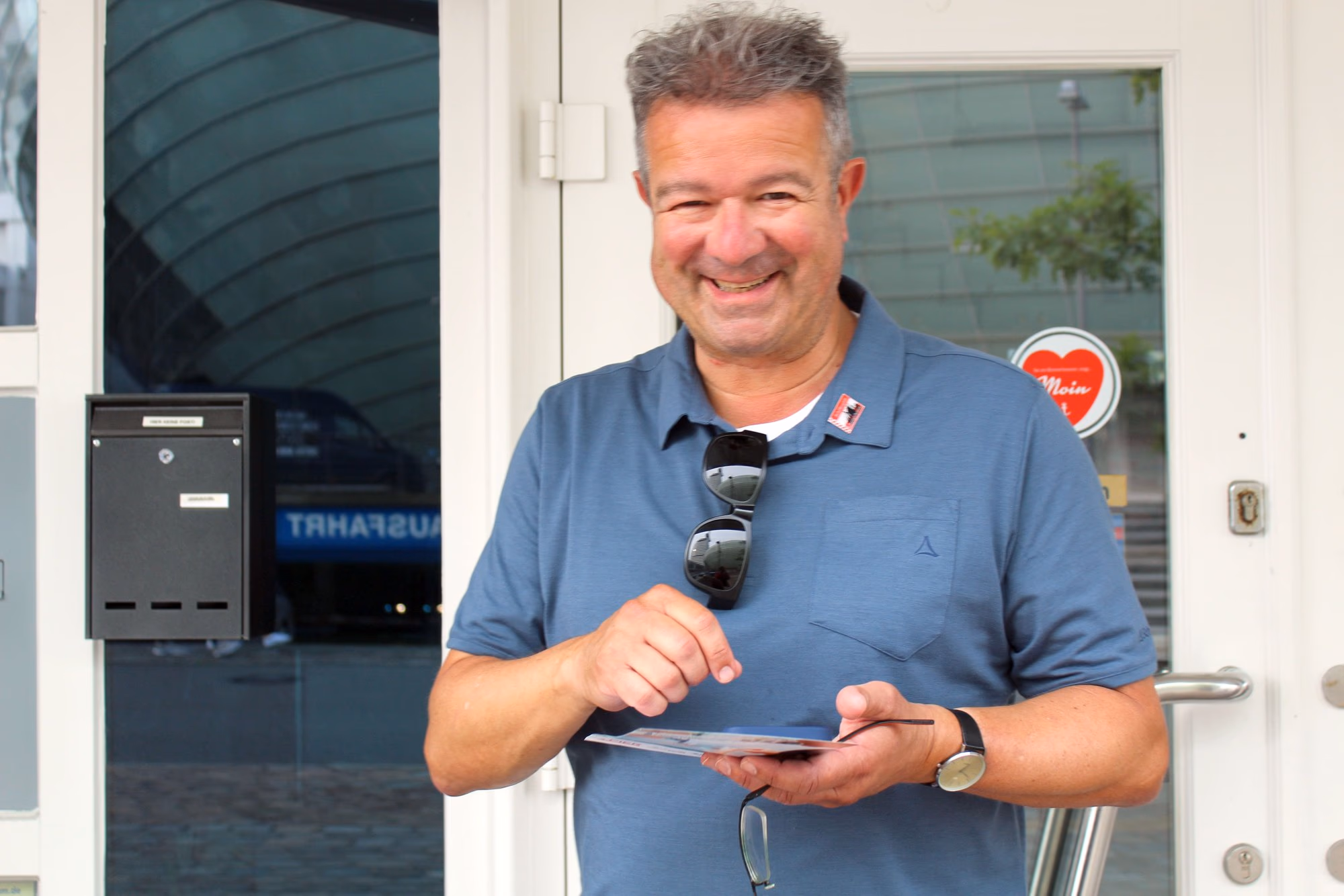 Manuel Andrack lächelt vor einer Glastür und hält eine Karte in der Hand