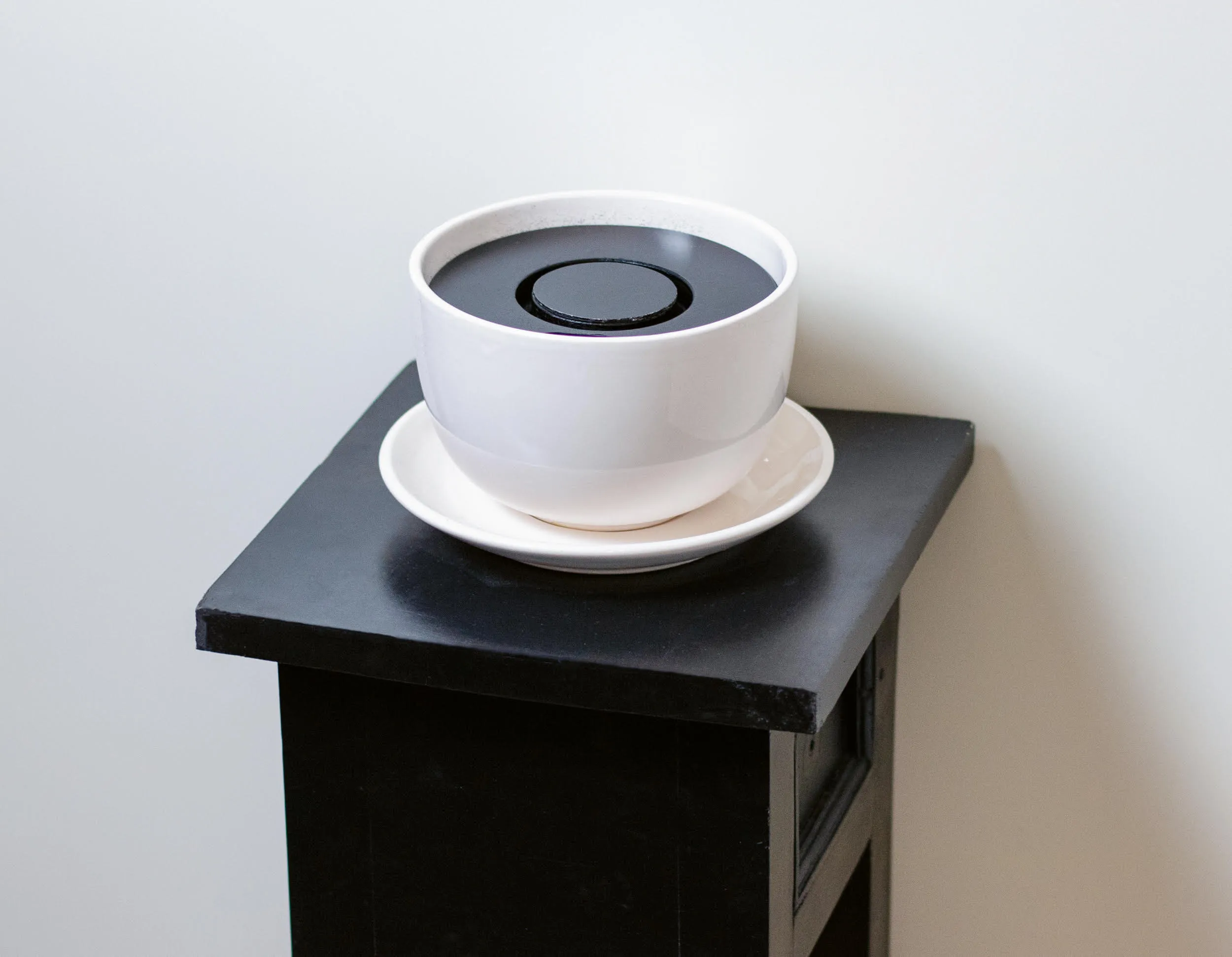 Der Cache ohne Getränk: eine weiße Tasse mit dunklem Einsatz auf einem schwarzen Sockel vor einer hellen Wand