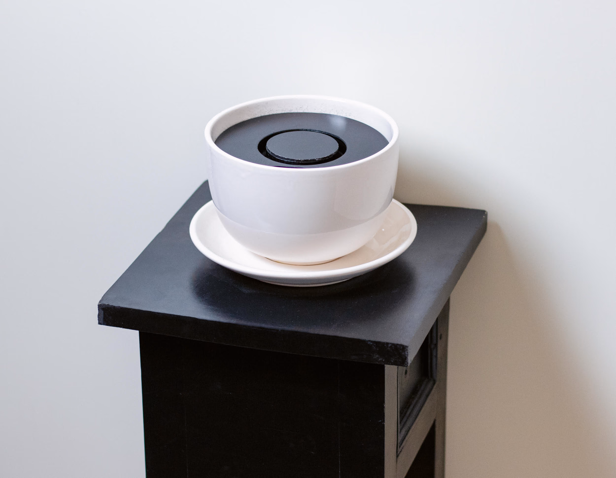 Der Cache ohne Getränk: eine weiße Tasse mit dunklem Einsatz auf einem schwarzen Sockel vor einer hellen Wand