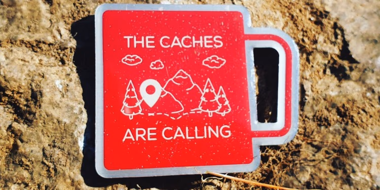 Rotes Schild in Form eines Bechers mit der Aufschrift „The Caches are calling“ auf felsigem Boden