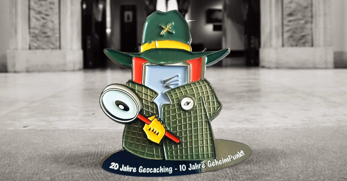 Geocoin als Figur mit grünem Hut, Mantel und Lupe, Aufschrift „20 Jahre Geocaching – 10 Jahre GeheimPunkt“, dahinter ein Museumssaal