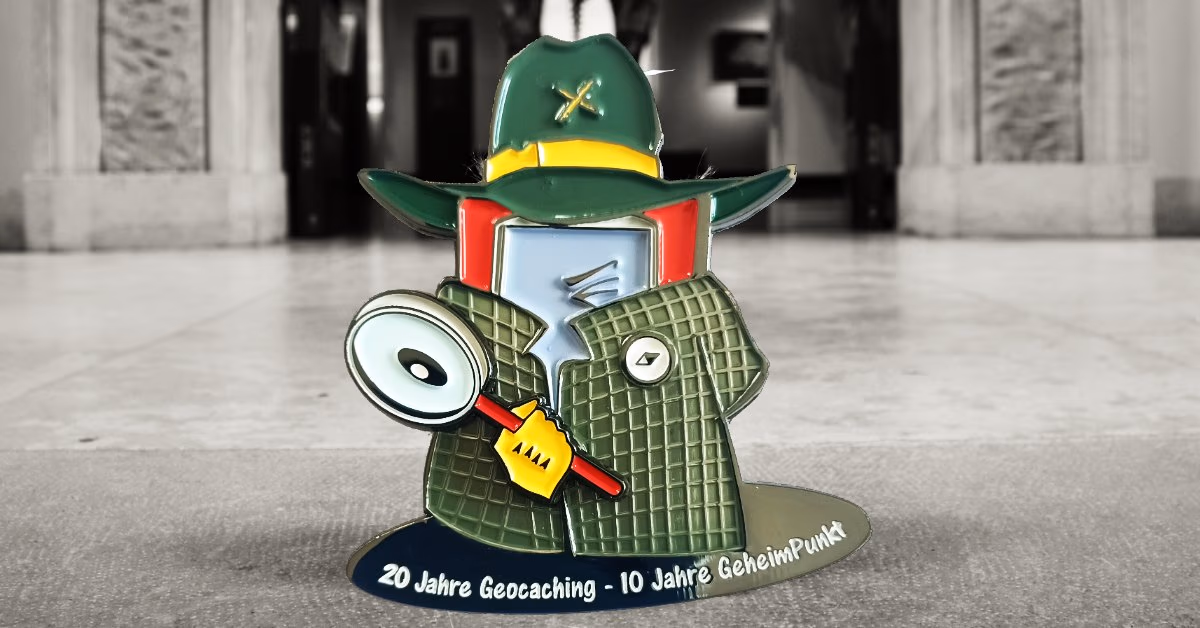 Geocoin als Figur mit grünem Hut, Mantel und Lupe, Aufschrift „20 Jahre Geocaching – 10 Jahre GeheimPunkt“, dahinter ein Museumssaal