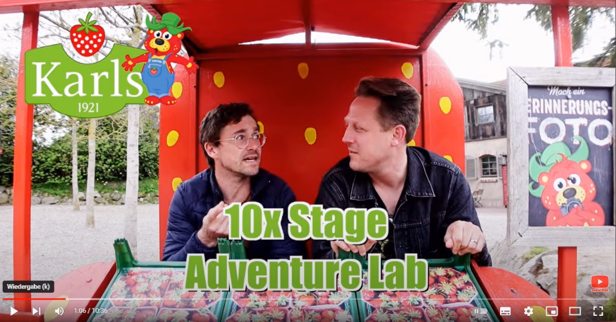 Standbild aus einem YouTube-Video: Daniel Pflieger und der Geocaching Vlogger in einem Erdbeer-Wagen bei Karls, Schriftzug „10x Stage Adventure Lab“