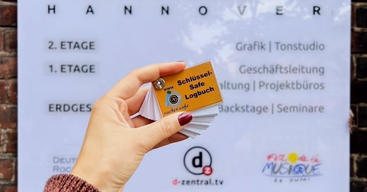 Hand hält ein kleines Logbuch mit der Aufschrift „Schlüssel-Safe Logbuch“ vor einer Etagentafel