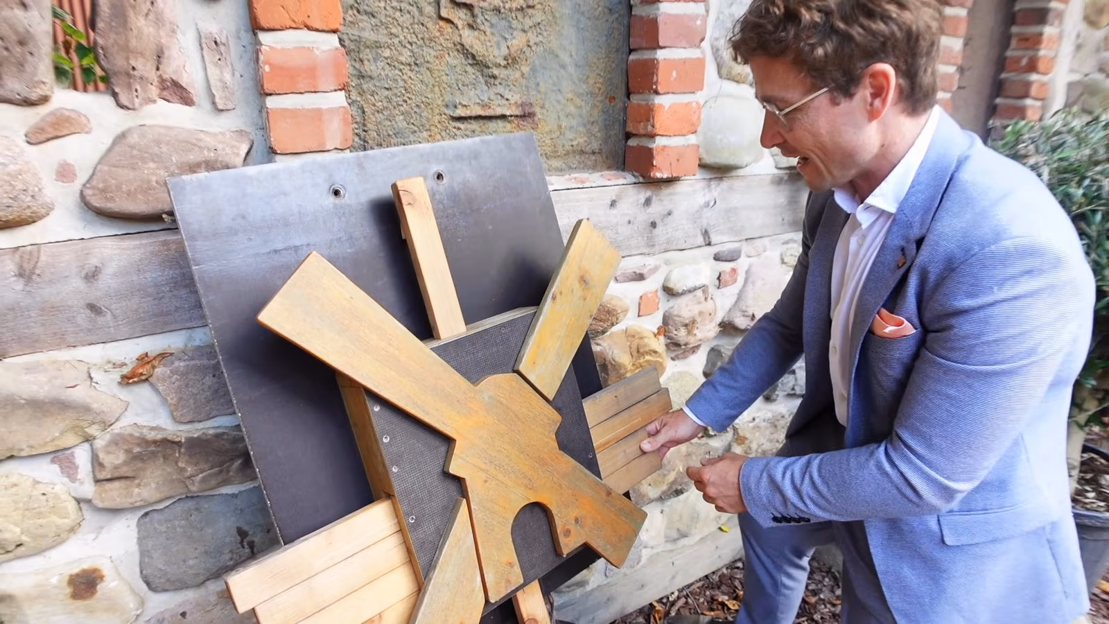 Daniel Pflieger im hellblauen Anzug an einem hölzernen Windmühlen-Rätsel vor einer Bruchsteinmauer