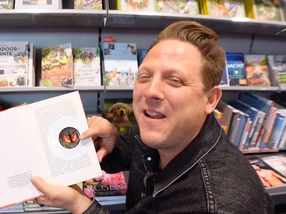 Der Geocaching Vlogger lacht in einer Hugendubel-Filiale zwischen den Regalen, in der Hand ein aufgeschlagenes Buch, in dem der nächste Hinweis steckt