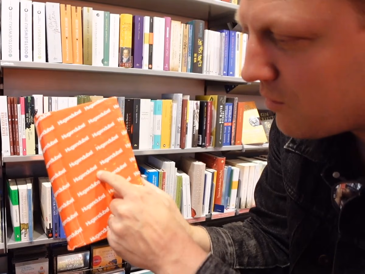 Zwischen den Regalen einer Hugendubel-Filiale steckt ein Hinweis in einem orangen Hugendubel-Umschlag