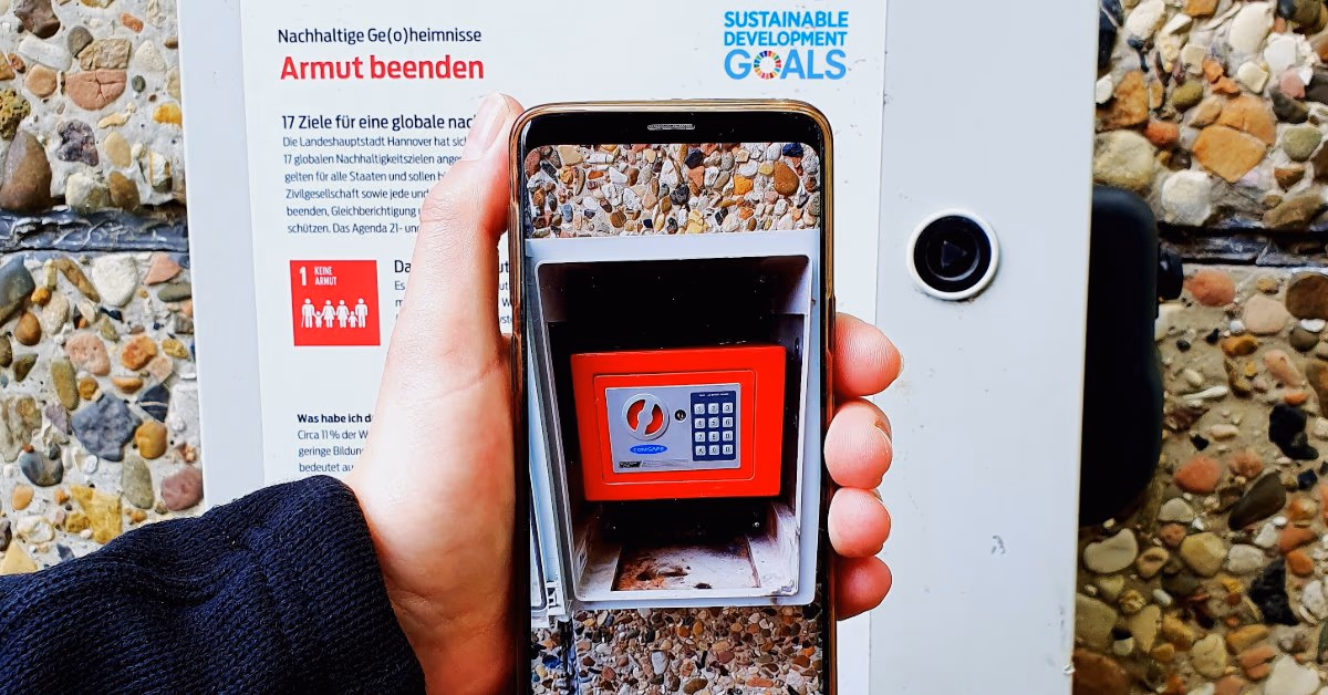 Hand hält ein Smartphone vor eine Tafel zum Nachhaltigkeitsziel „Armut beenden“, auf dem Display ein roter Tresor