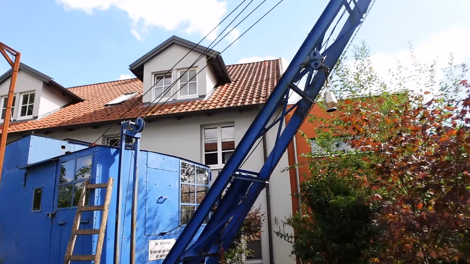 Blauer Seilbagger mit Gittermast vor einem Wohnhaus, am Ausleger hängt das Seil der Winde