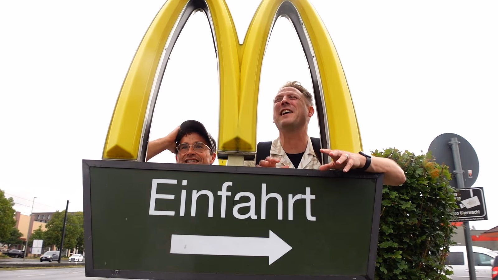 Daniel Pflieger und der Geocaching Vlogger lachen an der Einfahrt eines McDonald’s-Restaurants der Familie Ehmann, über ihnen das McDonald’s-Logo