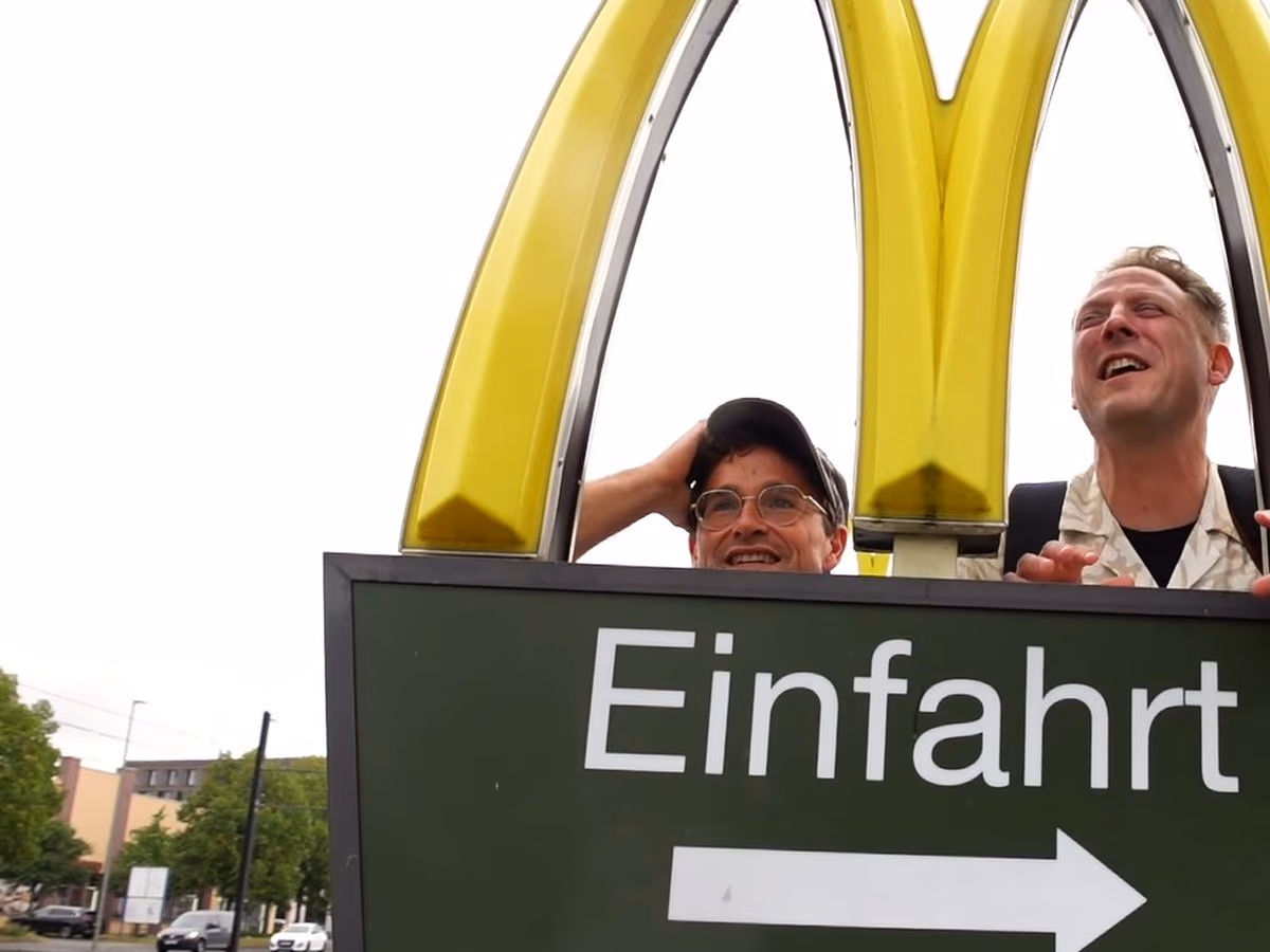 Daniel Pflieger und der Geocaching Vlogger lachen hinter dem Einfahrt-Schild eines McDonald’s-Restaurants, darüber das gelbe M