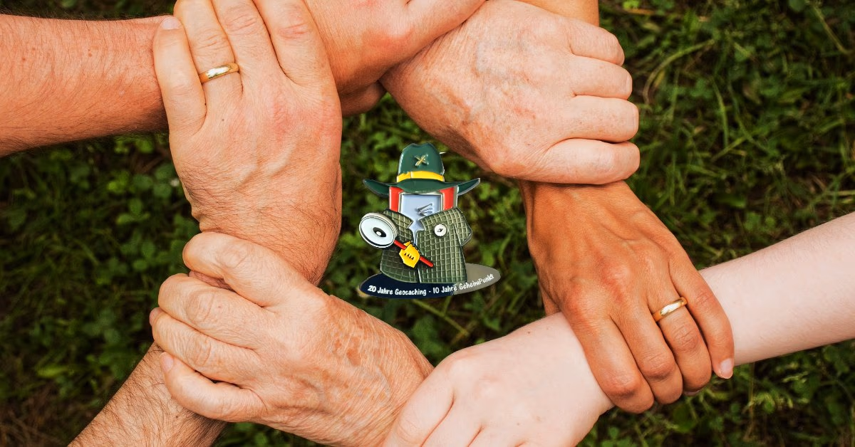 Vier Hände halten sich gegenseitig an den Handgelenken, in der Mitte eine Geocoin-Figur