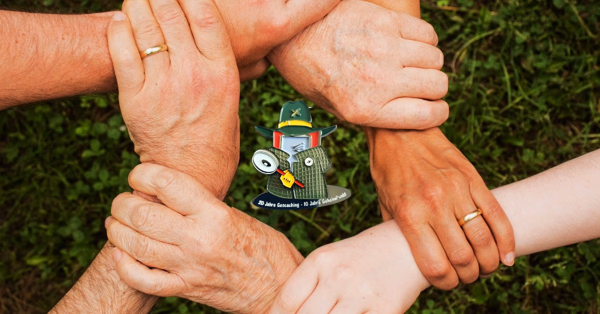 Vier Hände halten sich gegenseitig an den Handgelenken, in der Mitte eine Geocoin-Figur