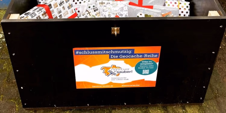 Große schwarze Holzkiste mit dem Aufkleber „#schlussmitschmutzig: Die Geocache-Reihe“