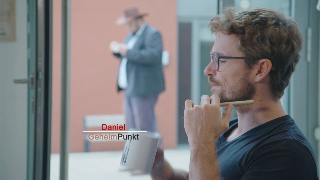 Daniel Pflieger im Profil an einer Glastür, im Hintergrund ein Mann mit Hut, der auf sein Smartphone schaut