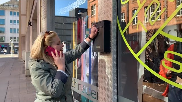 Eine Frau mit Smartphone am Ohr greift vor dem Schaufenster eines Outdoor-Geschäfts nach einem Kasten